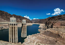 A large dam with two tall intake towers stands over a deep blue reservoir surrounded by rocky, arid terrain under a clear blue sky with scattered clouds. Description generated by AI