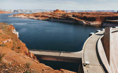 A large concrete dam spans a deep blue reservoir surrounded by red rocky cliffs under a partly cloudy sky. Description generated by AI